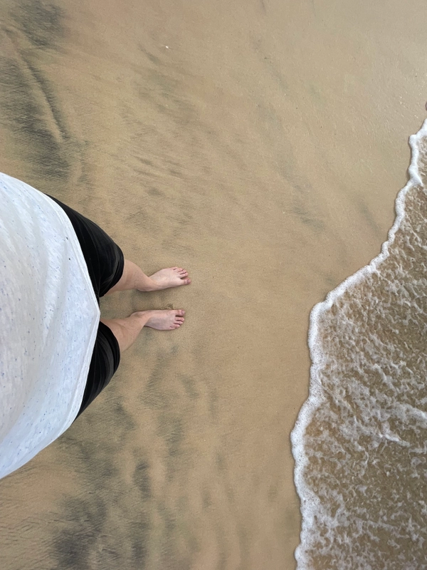 A person is standing on the beach near the water. Only the lower half of the person's body is visible. He is wearing short dark trousers, a light-coloured T-shirt and no shoes. The sand is dark and the water is slightly foamy.