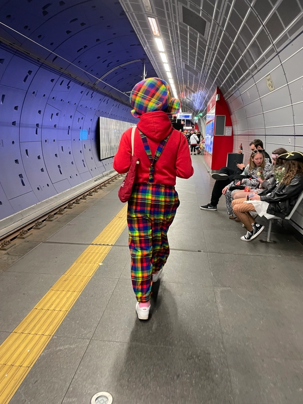A male person is walking along an underground station. The person is dressed as a clown in a carnival costume with colourful trousers, a colourful hat and a red jumper. In the background, people in fancy dress are sitting on a bench.