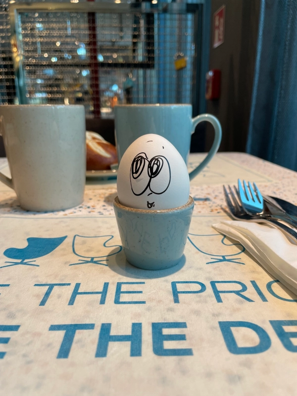 A light-coloured egg lies in a blue egg cup on a breakfast table. A face with large eyes and a nose has been drawn on the egg with a dark, thin pencil. In the background are blue cups, cutlery for two people and bread rolls.