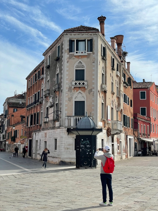 In the background, an old, ochre-coloured corner house on an empty, cobbled street. The neighbouring houses are painted red and some have long chimneys. The sky is light blue with a few clouds. In the foreground, a man with a bright red backpack is taking a photo of the corner house.